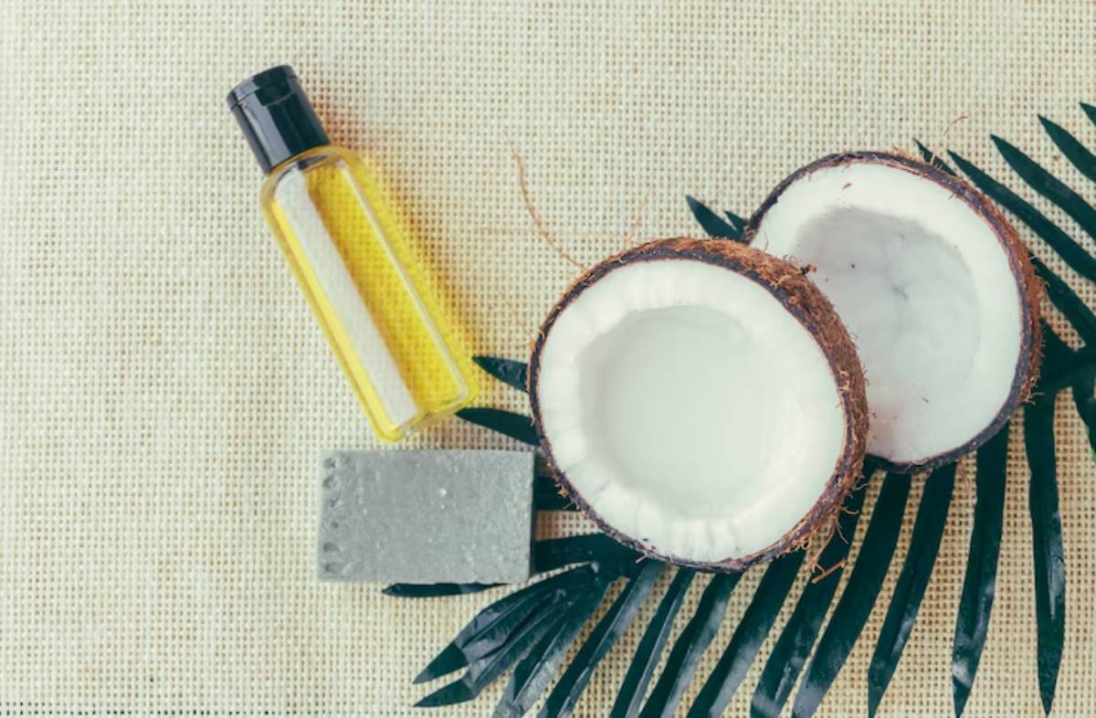 A bottle of yellow oil next to halved coconuts and a gray soap bar, arranged on a textured beige surface with green leaves.