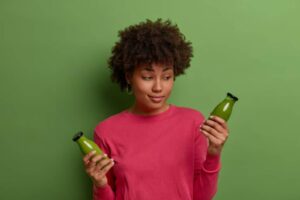 A person holds two small green juice bottles, smiling against a vibrant green background, wearing a pink sweatshirt and earrings.