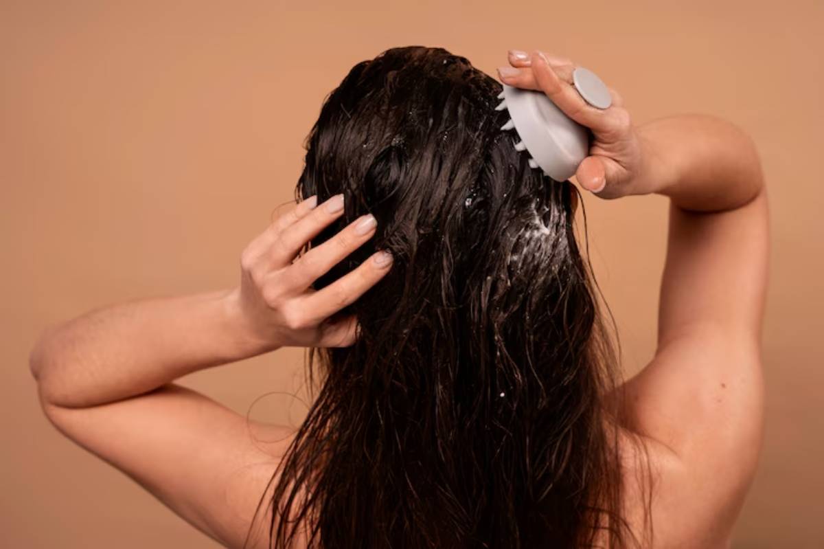 A woman applies hair product to her wet, long hair using a massaging tool, against a soft, neutral-toned background.