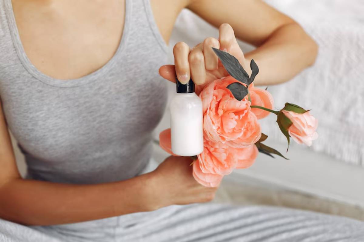 A person in a gray tank top holds a small bottle of lotion next to a bouquet of soft pink roses.