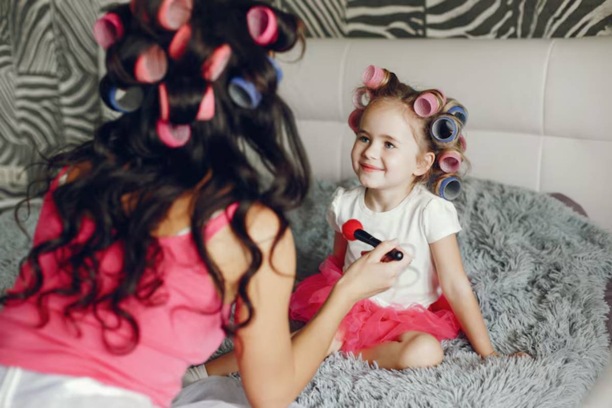 A woman prepares a young girl for a makeover, using a red brush on her face, both adorned with colorful hair rollers.