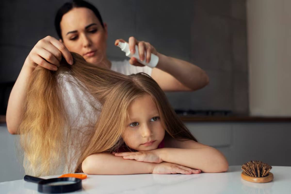 A person is applying hair spray to a child's long hair while the child rests their head on their arms at a table.