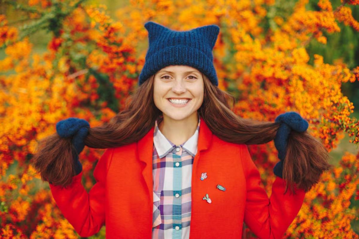 A person in a bright orange jacket and blue hat stands in front of vibrant orange flowers, holding their long brown hair.
