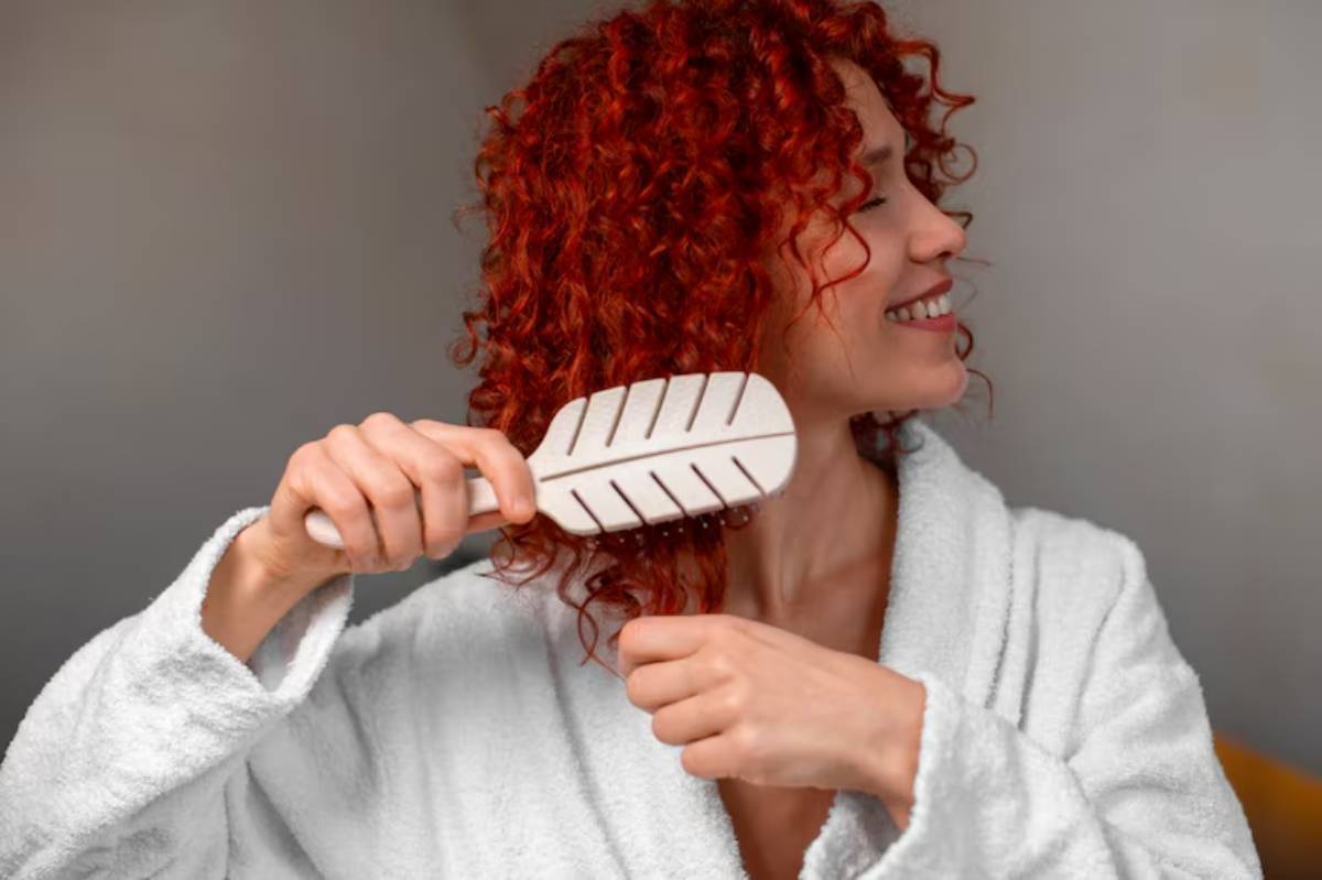 A person with vibrant red curly hair wearing a white robe brushes their hair in a softly lit indoor setting.