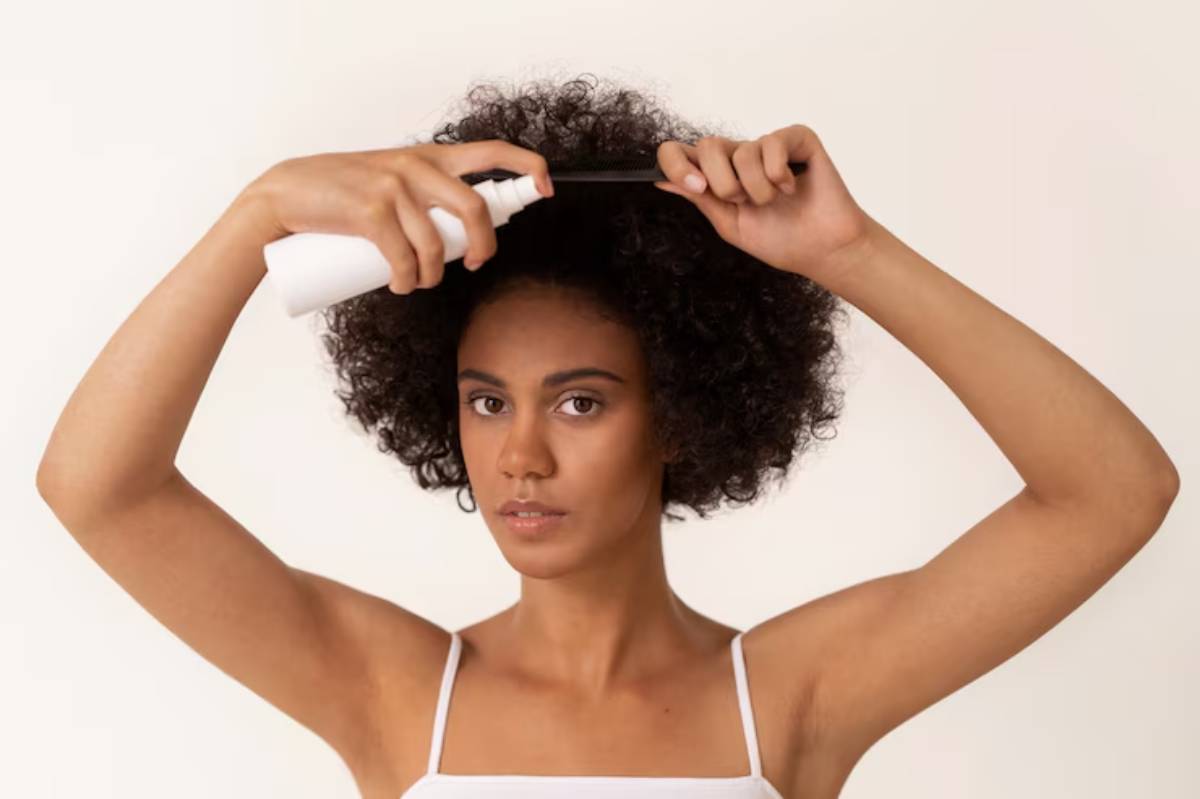 A person with curly hair spray styling product, holding a comb, against a neutral background.