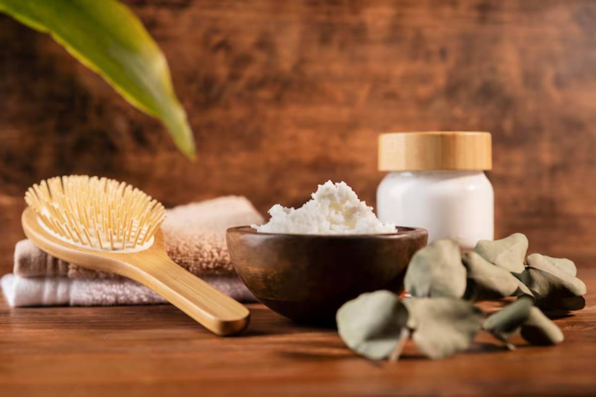 A wooden bowl filled with cream, a bamboo hairbrush, white towels, and eucalyptus leaves on a wooden surface. Natural skincare setup.