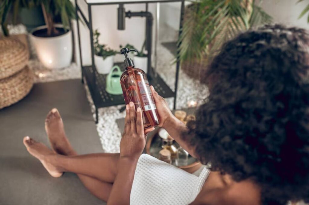 A woman with curly hair examines a brown bottle in a serene, plant-filled setting, sitting on the floor in a white robe.