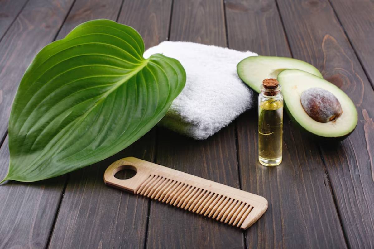 A wooden comb next to a towel, avocado halves, a bottle of oil, and a large green leaf on a wooden surface.