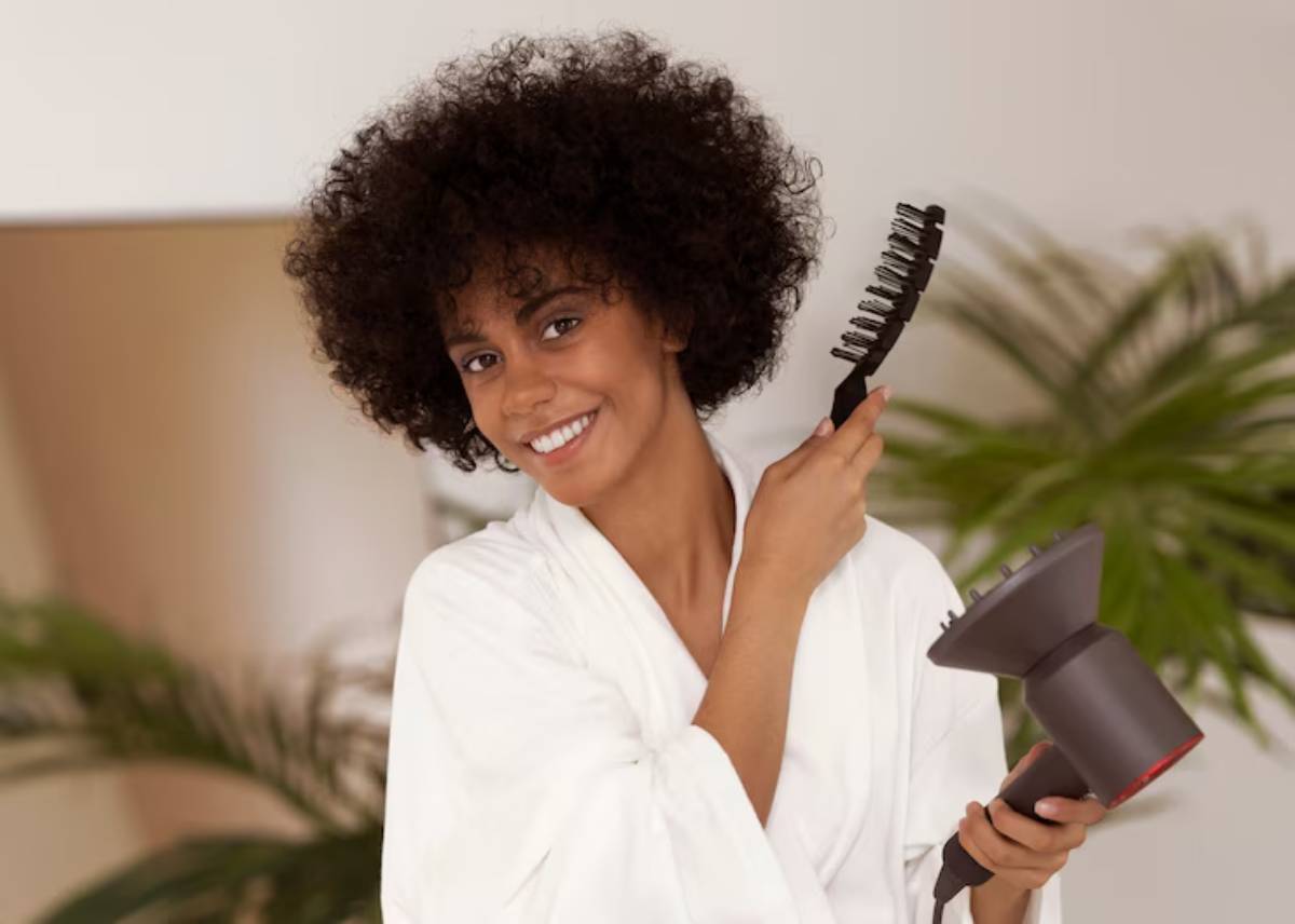 A person in a white robe holds a hairdryer and a comb, preparing to style their curly hair, with tropical plants in the background.