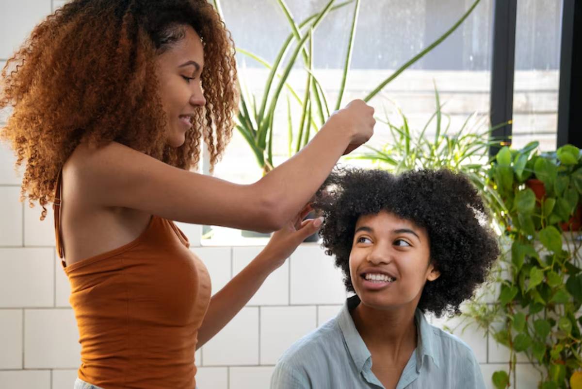 A person with curly hair receives a hairstyle treatment from another, surrounded by greenery in a bright indoor space.
