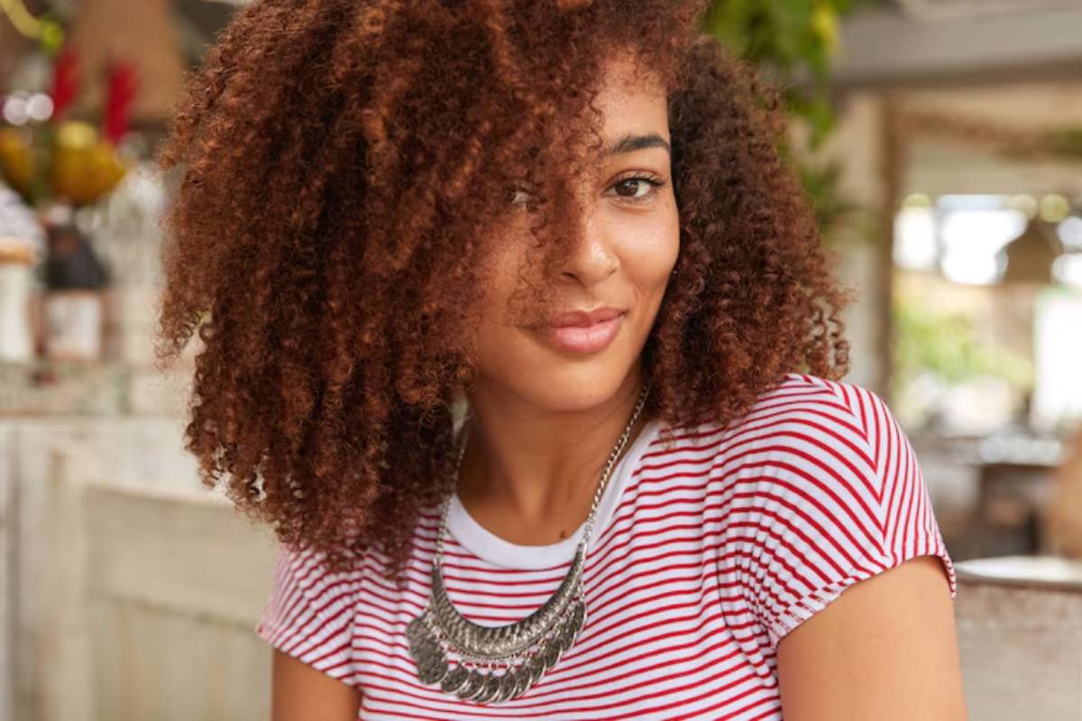 A close-up of a woman with curly, voluminous hair wearing a red and white striped shirt and a layered silver necklace.