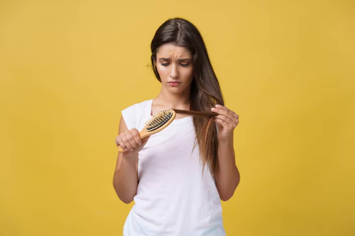A person holds a hairbrush in one hand and a strand of long, brown hair in the other, set against a bright yellow background.