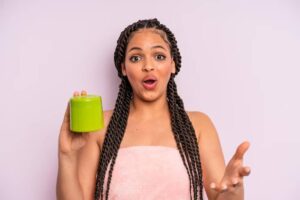 A person with long braided hair holds a green moisturizing cream, wearing a pink towel against a soft purple backdrop.