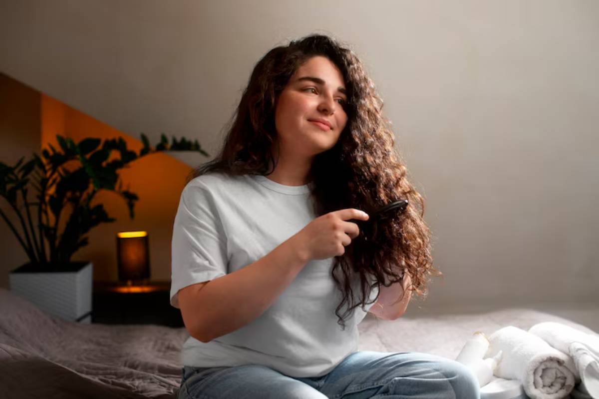 A person with long, curly hair sits on a bed, brushing their hair in a cozy, softly-lit room with plants nearby.