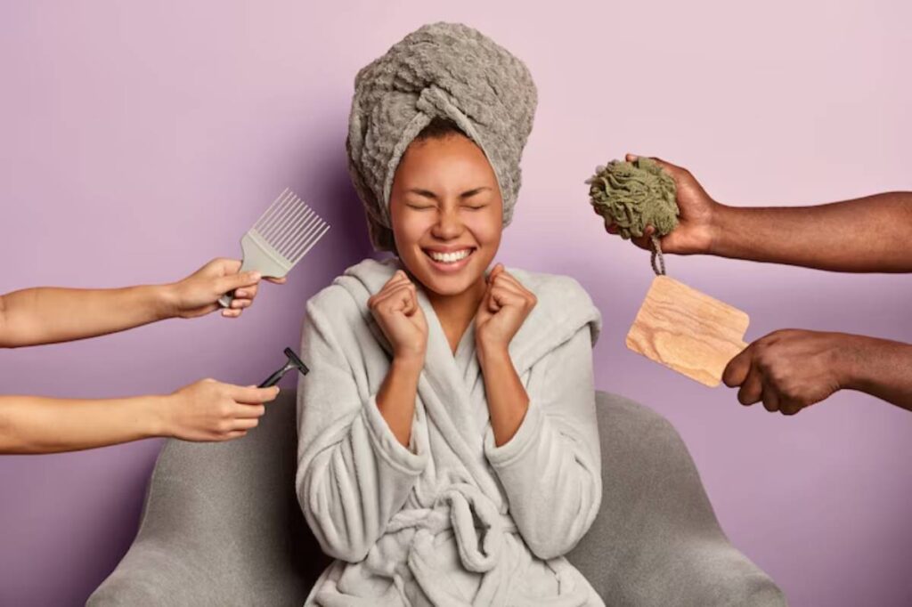 A person in a cozy bathrobe is surrounded by grooming tools, including a comb, razor, exfoliating sponge, and wooden board, against a pink backdrop.