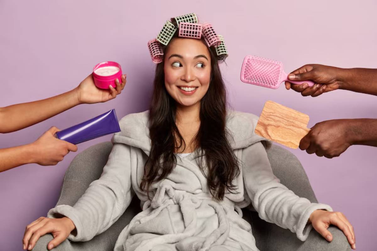 A person in a cozy robe sits in a chair, surrounded by various hair products and styling tools, under colorful hair rollers.