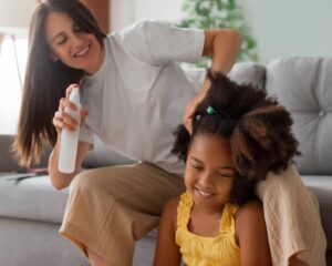 A woman styles a child's curly hair using a spray bottle, both seated comfortably on a sofa in a bright, modern living room.