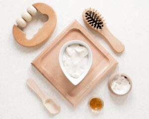 A flat lay of wooden skincare tools and products, including a cream in a bowl, a brush, honey, and a scoop on a light surface.