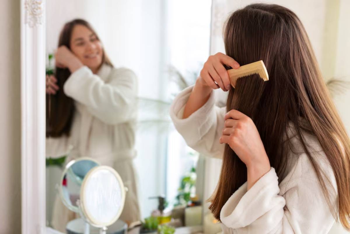 A person wearing a white robe combs their long hair in front of a mirror, with plants and a vanity in the background.