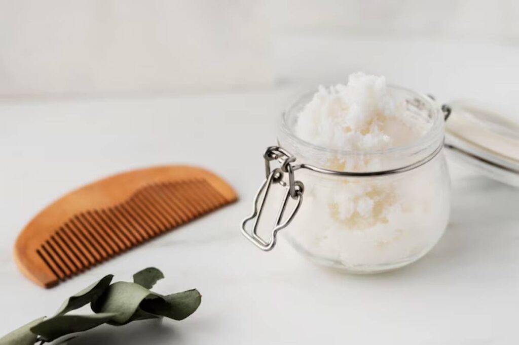 A glass jar filled with fluffy hair cream, a wooden comb, and a sprig of eucalyptus on a soft white surface.