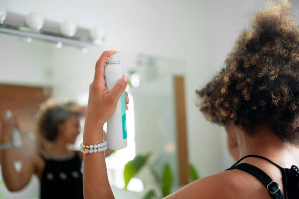 A person holds a spray can in a bright bathroom, preparing to style their curly hair in front of a mirror.