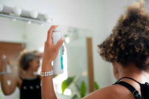 A person holds a spray can in a bright bathroom, preparing to style their curly hair in front of a mirror.