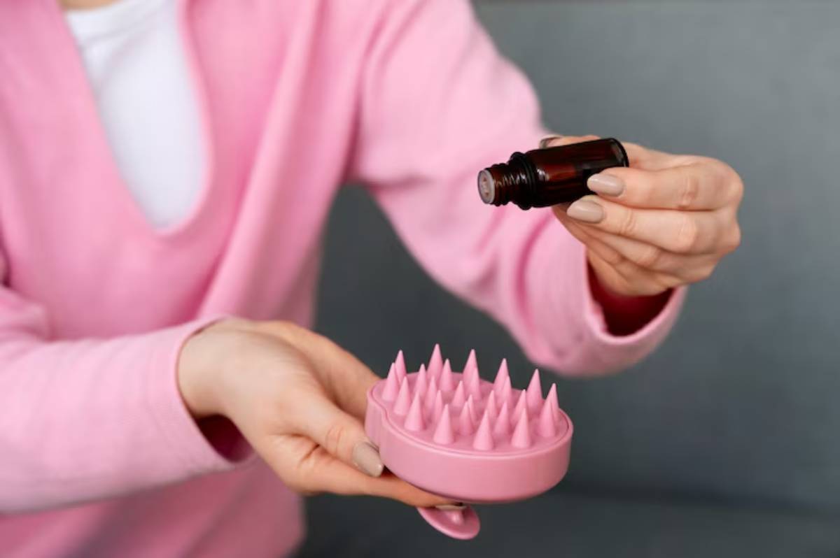 A person in a pink sweater holds a bottle of oil above a pink massager with spiky tips, preparing for a hair massage.