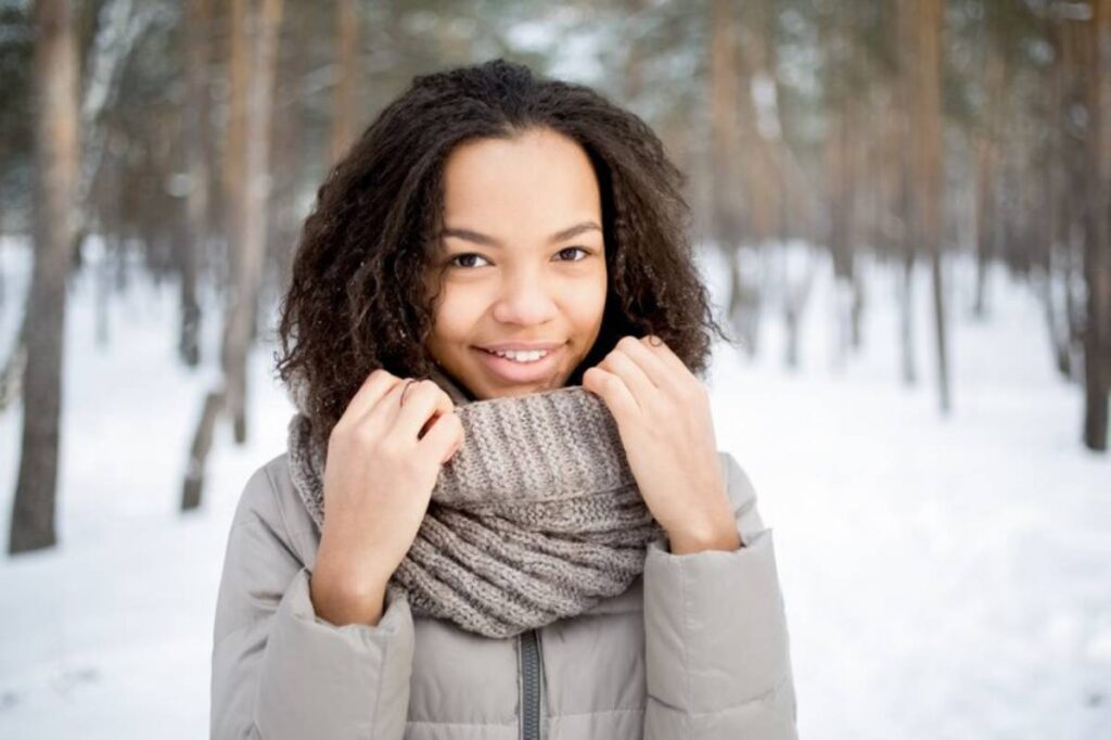 A person in a cozy gray jacket and knit scarf stands outdoors in a snowy forest, surrounded by tall trees.
