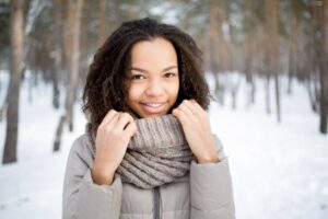 A person in a cozy gray jacket and knit scarf stands outdoors in a snowy forest, surrounded by tall trees.