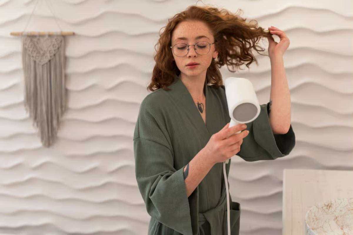 A person in a green robe holds a hairdryer while styling their curly hair against a textured cream wall with hanging decor.