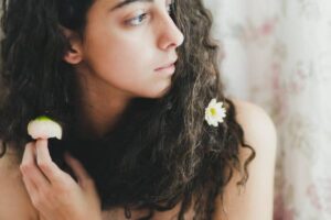 A person with long, curly hair adorned with a white flower holds a pink bud delicately between their fingers. Soft floral backdrop.