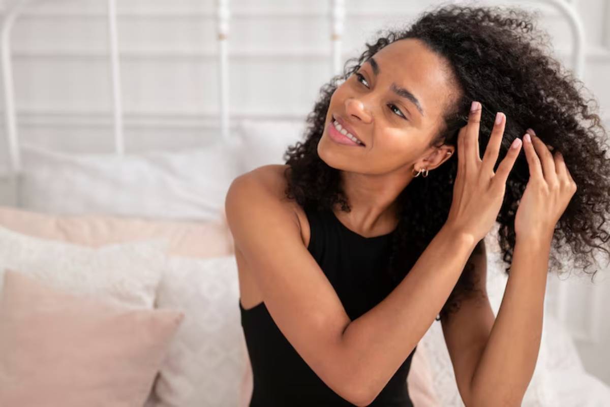A woman with curly hair elegantly styles her hair while sitting on a bed adorned with soft, neutral-colored pillows.