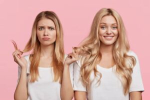 Two women with long hair, one straight and one wavy, both wearing white t-shirts, stand against a pink background.