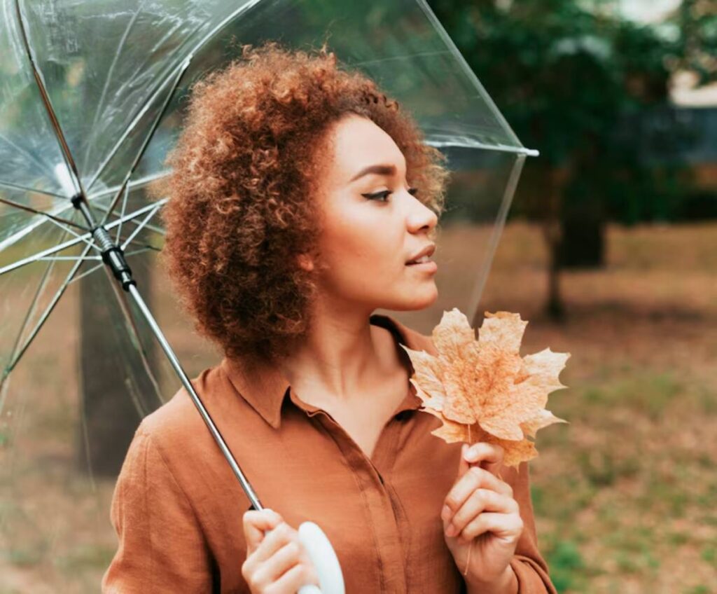 A woman holds a large, dry leaf while standing under a clear umbrella in a park with autumn foliage in the background.