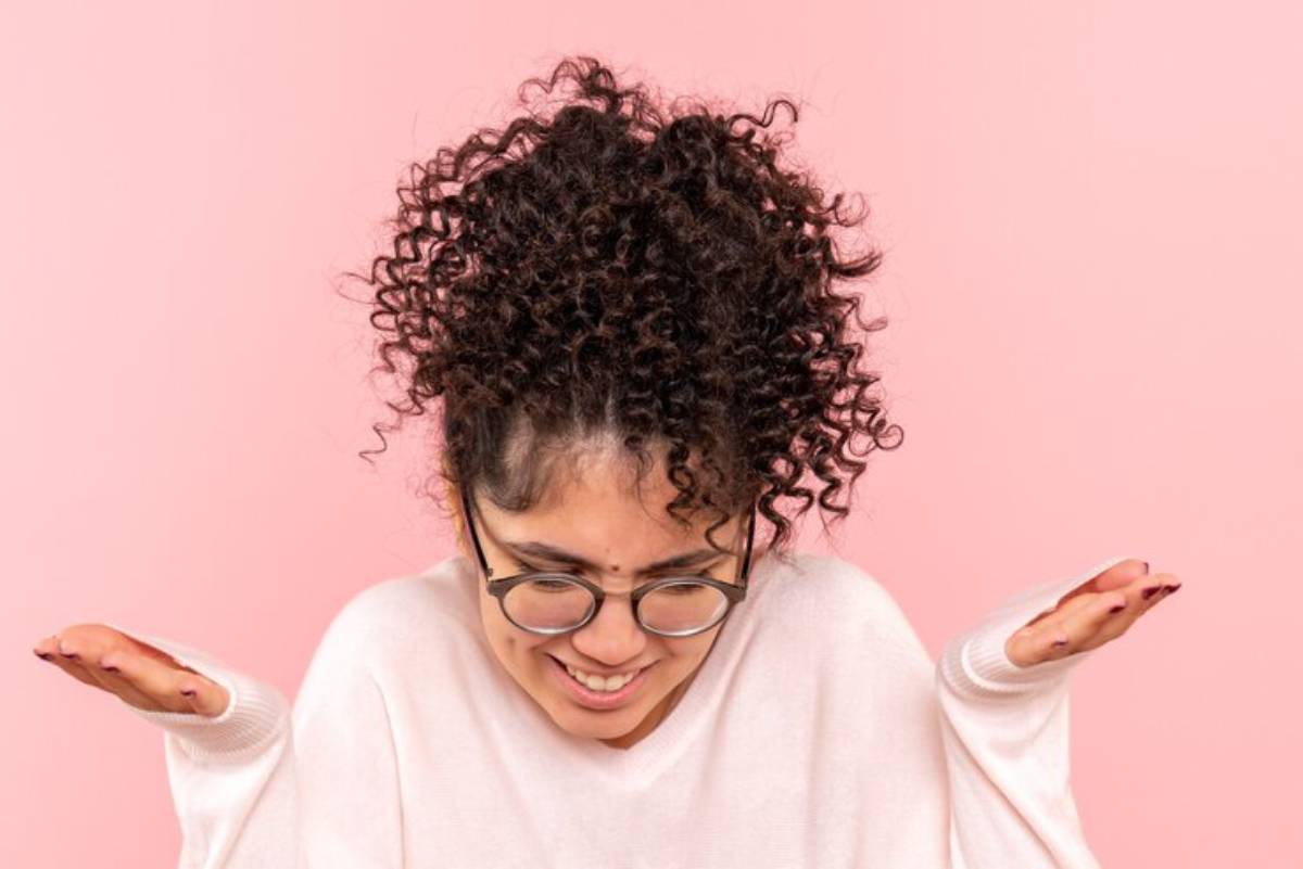 A person with curly, dark hair wearing a light sweater, holding their hands up with a confused expression against a pink background.