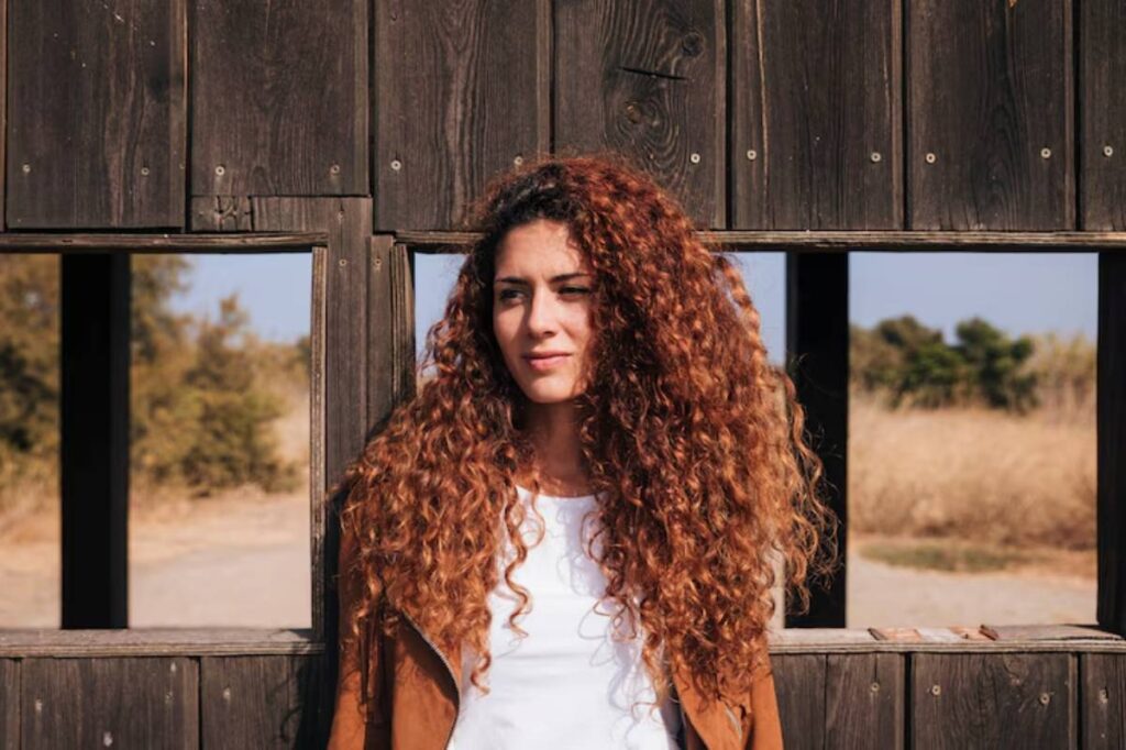 A person with long, curly hair stands in front of a wooden structure with open windows, surrounded by tall grass and nature.