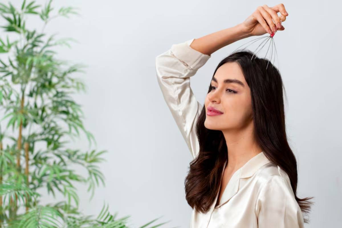 A woman in a silky blouse playfully holds a wire hair massager above her head, surrounded by lush green foliage.