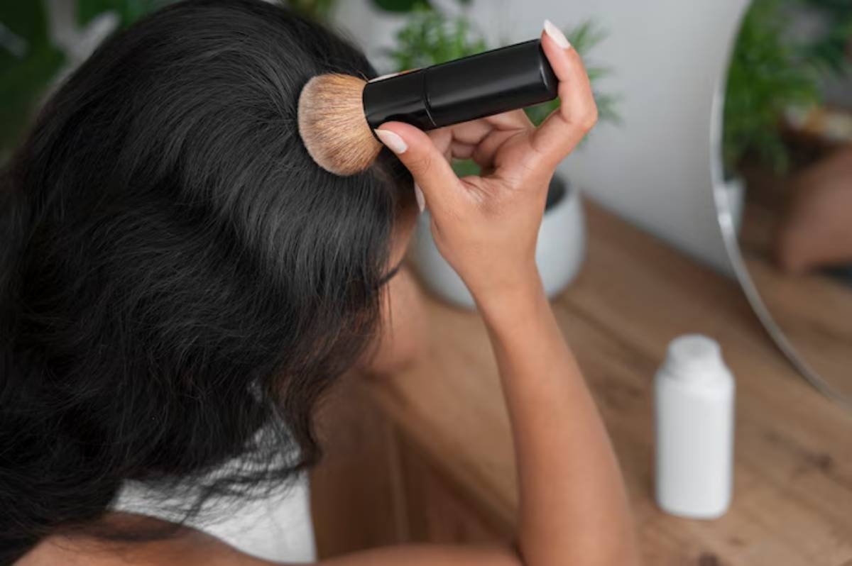 A woman applies makeup with a brush to her dark hair, seated at a wooden table with a mirror and a white bottle nearby.