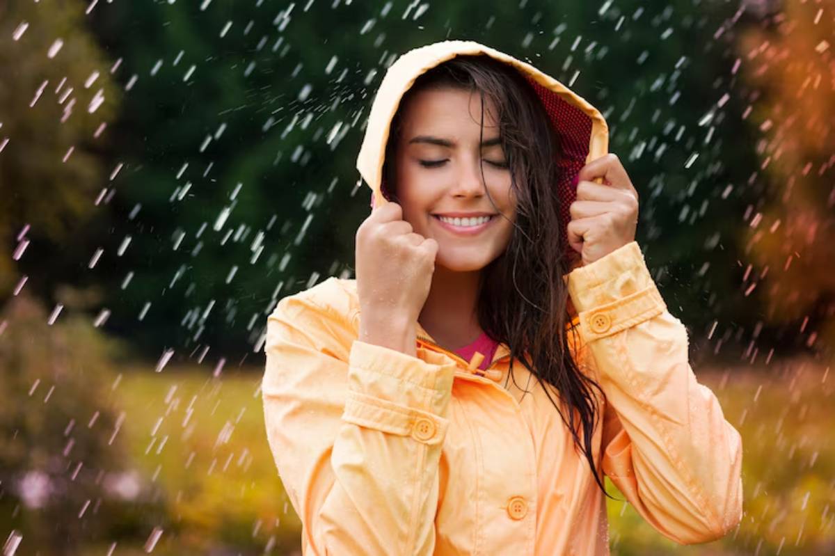 A person in a bright orange raincoat stands in the rain, holding the hood, with colorful leaves in the blurred background.