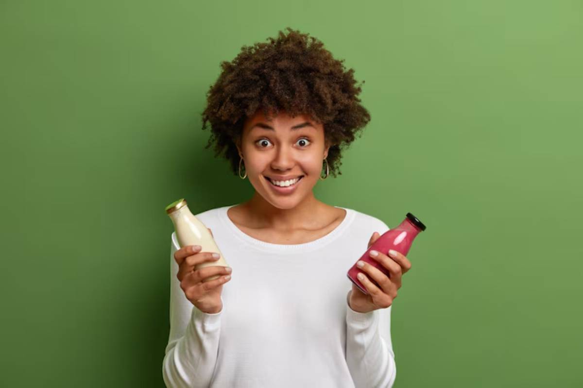 A person with curly hair holds two glass bottles, one filled with creamy white liquid and the other with a vibrant red drink, against a green backdrop.