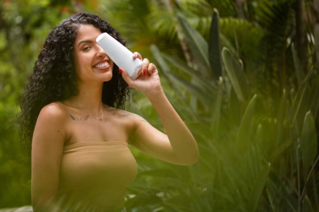 A woman with curly hair holds a white drink container while surrounded by lush green plants, enjoying a sunny outdoor moment.