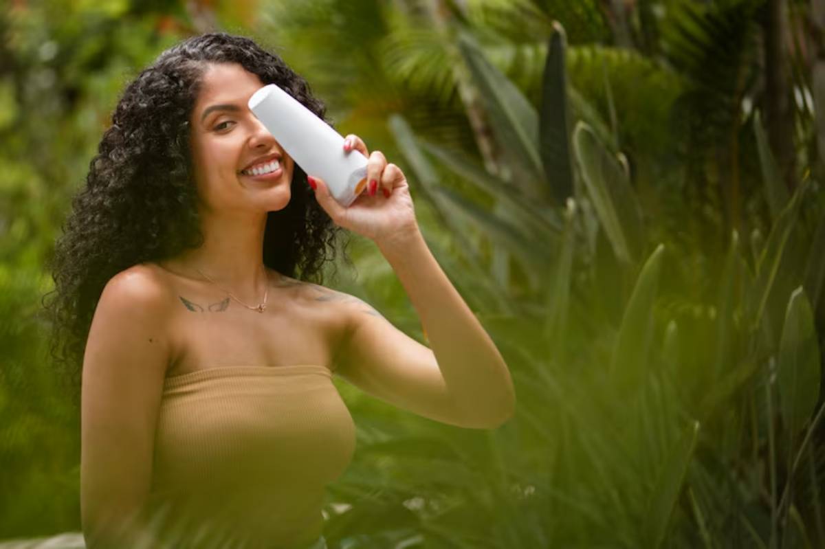 A woman with curly hair holds a white drink container while surrounded by lush green plants, enjoying a sunny outdoor moment.