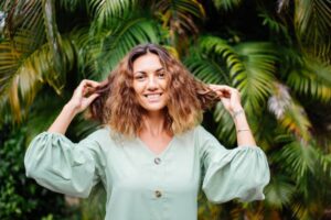 A woman with wavy hair, wearing a mint green blouse, poses playfully with her hair against a lush, tropical background.