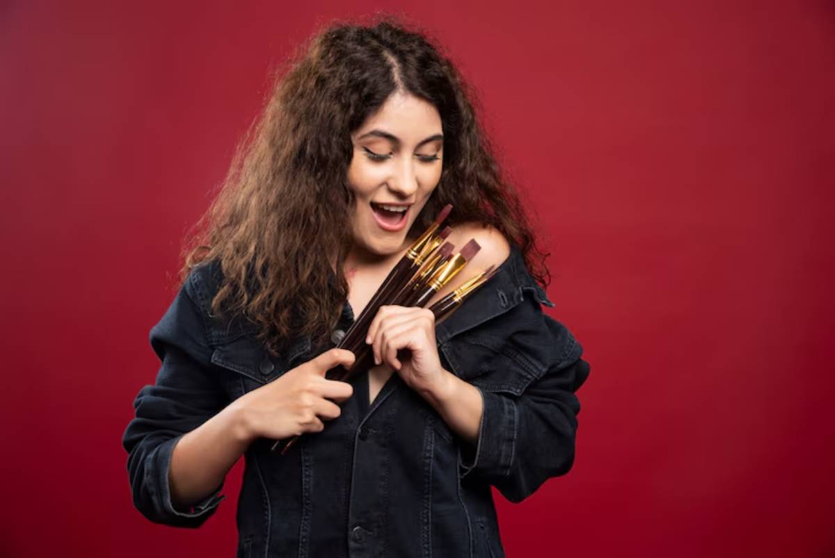 A woman in a black denim jacket stands against a red background, holding several makeup brushes.