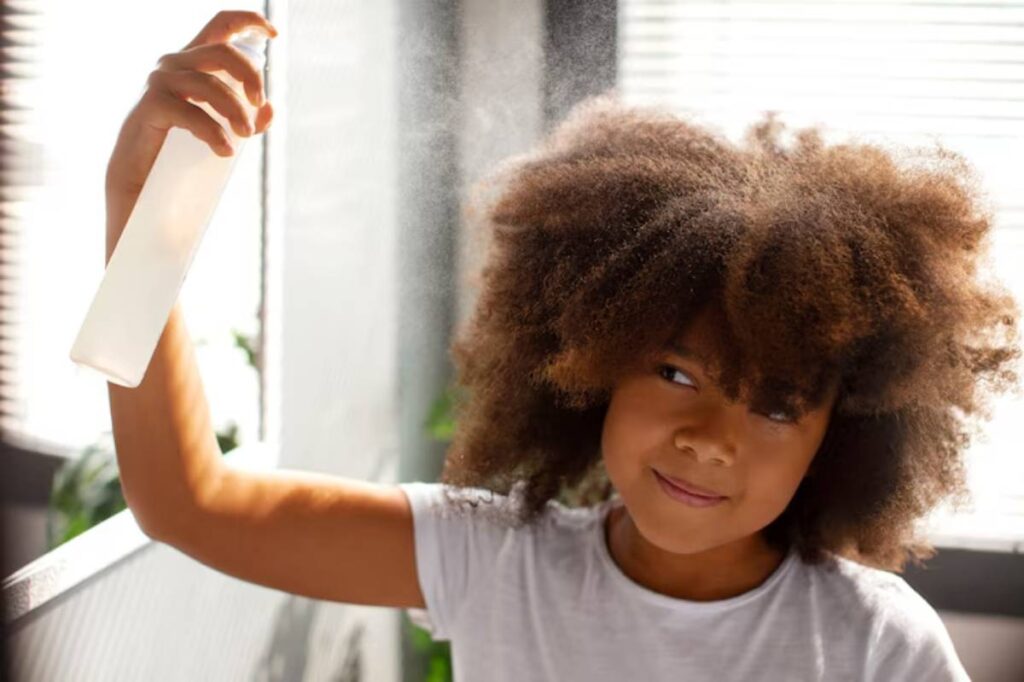 A person with voluminous, textured hair holds a spray bottle, misting their hair in bright sunlight filtering through blinds.