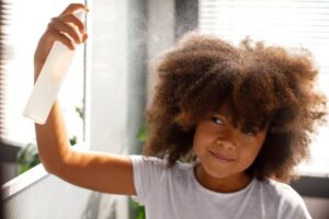 A person with voluminous, textured hair holds a spray bottle, misting their hair in bright sunlight filtering through blinds.
