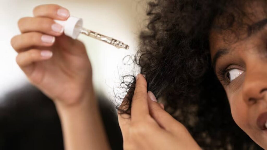 A woman applies hair serum from a dropper to her curly hair, focusing on nourishing and enhancing its texture.
