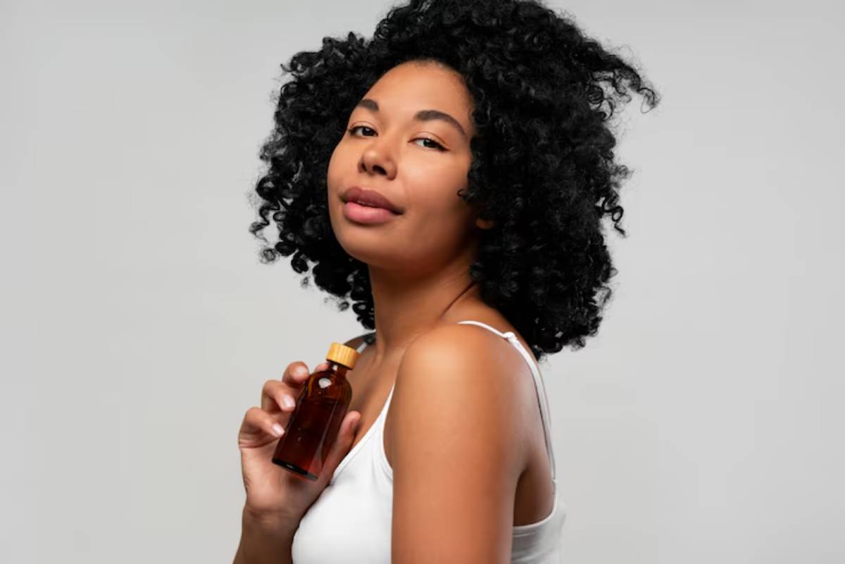 A woman with curly hair holds a small amber bottle with a wooden cap, wearing a white tank top against a light gray background.