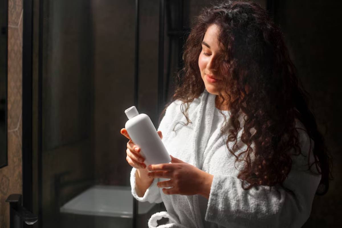 A person in a white bathrobe holds a white shampoo bottle, standing in a softly lit bathroom near a shower.