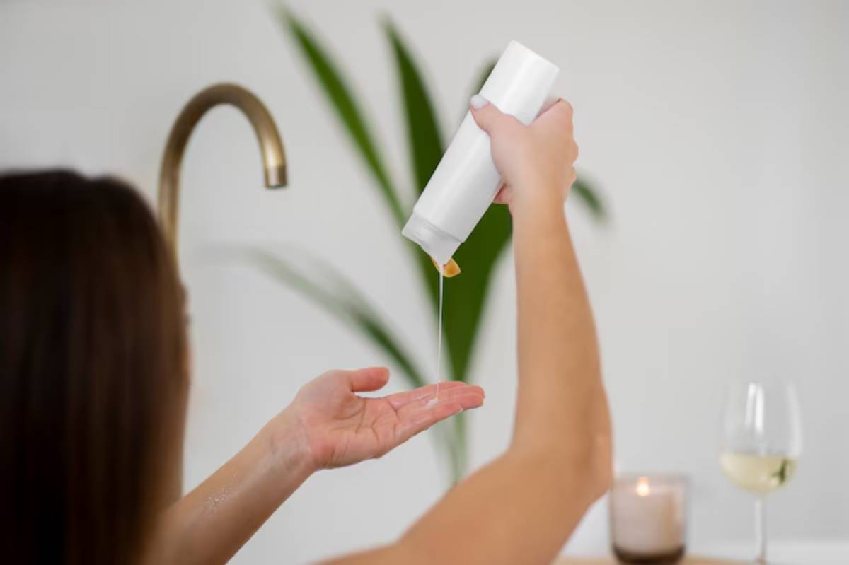 A person pours oil from a white bottle into their palm, with a candle and wine glass in the background and green plants nearby.